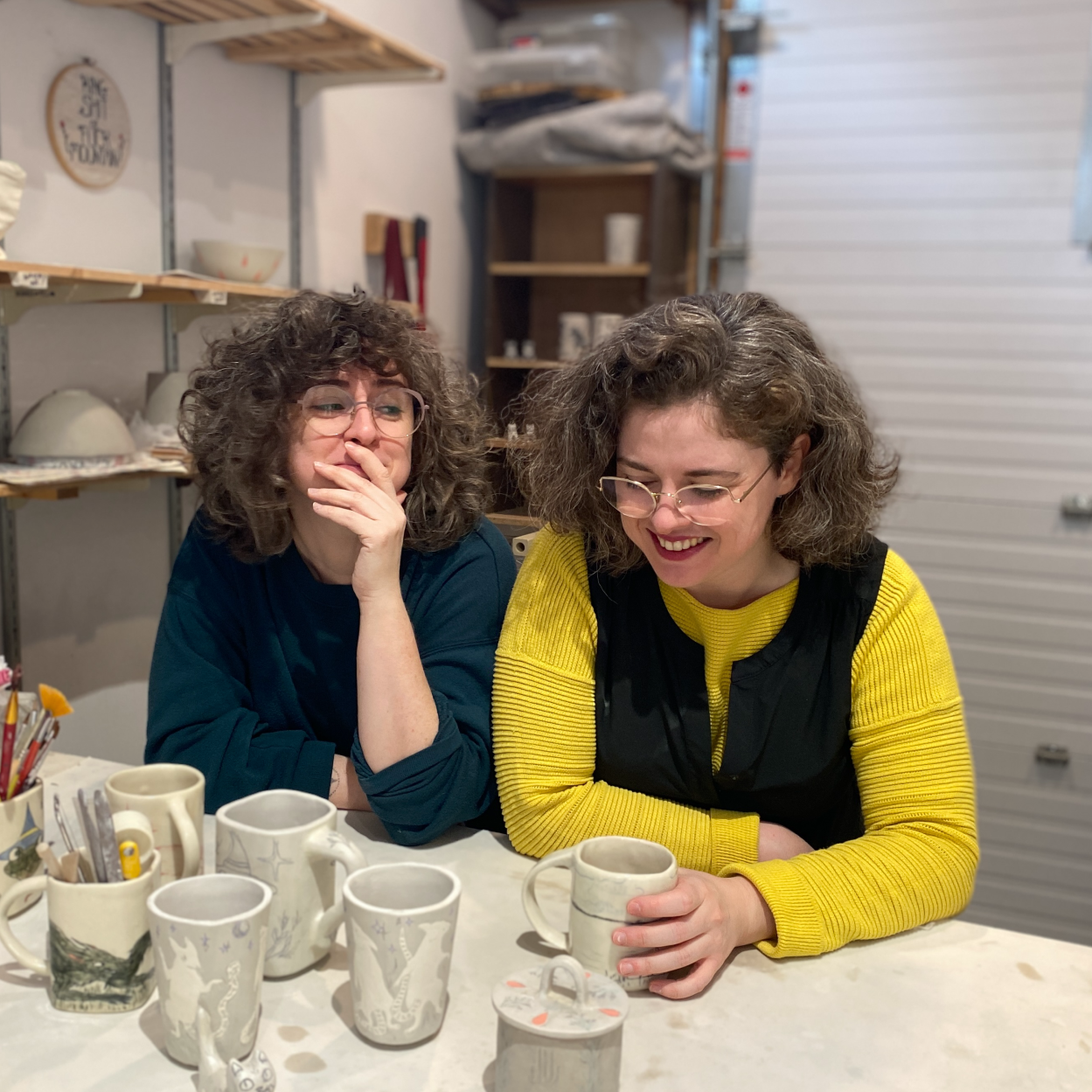 A photograph of Cass and Maia. Cass is covering her mouth and laughing while looking at Maia, who smiles while holding a handmade mug.